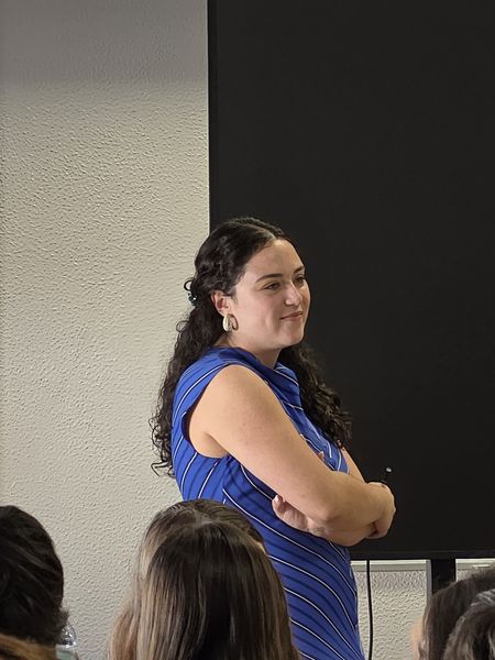 Mulher de vestido azul às riscas, de pé junto ao ecrã, a ouvir a conversa